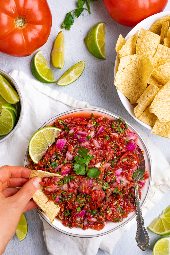 A bowl of easy fermented salsa with a hand dipping a tortilla chip in to the salsa. A spoon is also in the salsa. A bowl of tortilla chips is off to the side, along with a bowl of limes, and a few scattered tomatoes.