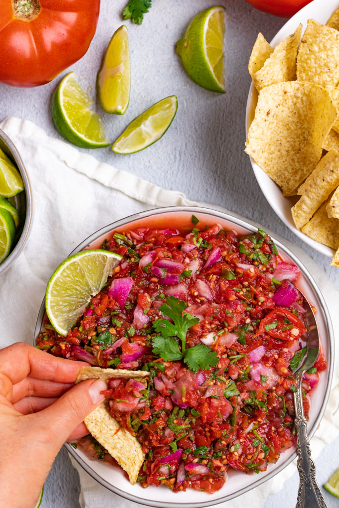 Easy fermented salsa in a bowl with a hand dipping a tortilla chip into it. There is a tomato scattered in the background, along with limes, and tortilla chips.