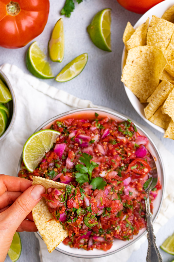 A closeup shot of the fermented salsa being with a hand holding a chip with salsa on it closer to the camera. Limes are scattered in the background, along with chips, and tomatoes.