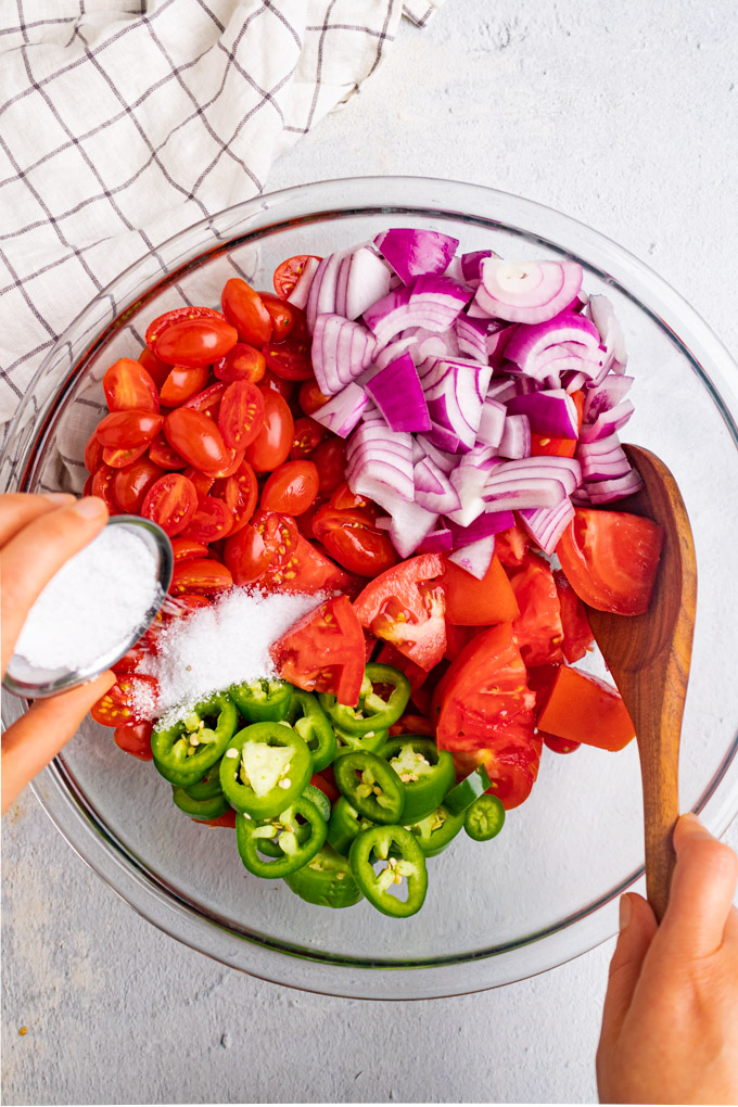 A bowl of cut up tomatoes, jalapenos, and onion. A hand is pouring a small bowl of salt into the tomato mixture. A wooden spoon is being held by another hand.