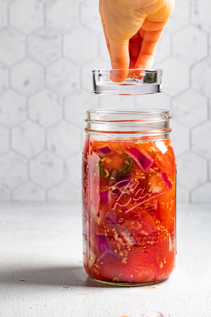 A mason jar is full of tomatoes with a lot of liquid. A glass fermenting weight is being held by a hand, above the mason jar.