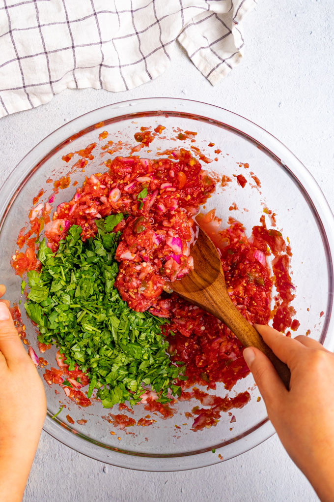 A large bowl of fermented salsa wit a pile of cilantro being stirred in by a hand holding a wooden spoon. 