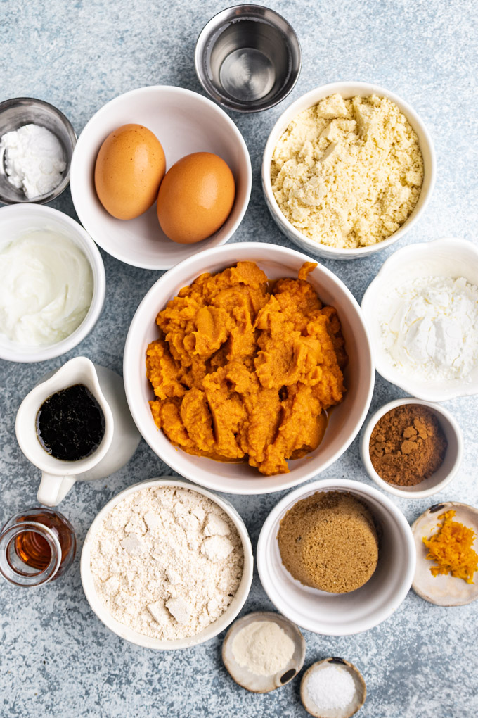 Ingredients for pumpkin bread are in bowls, on a light blue background. Pumpkin puree, eggs, flour, sugar, maple syrup, yogurt, pumpkin pie spice, etc.