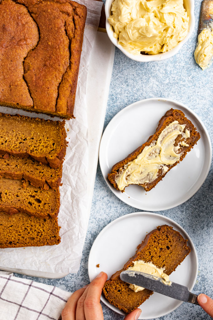 Healthy pumpkin bread is on a light blue background with the loaf of bread off to the side. It is sliced up, on a white marble board. Two slices of pumpkin bread are off the the side, on plates. One has butter spread onto it while the other has a hands, and knife spreading butter onto it.
