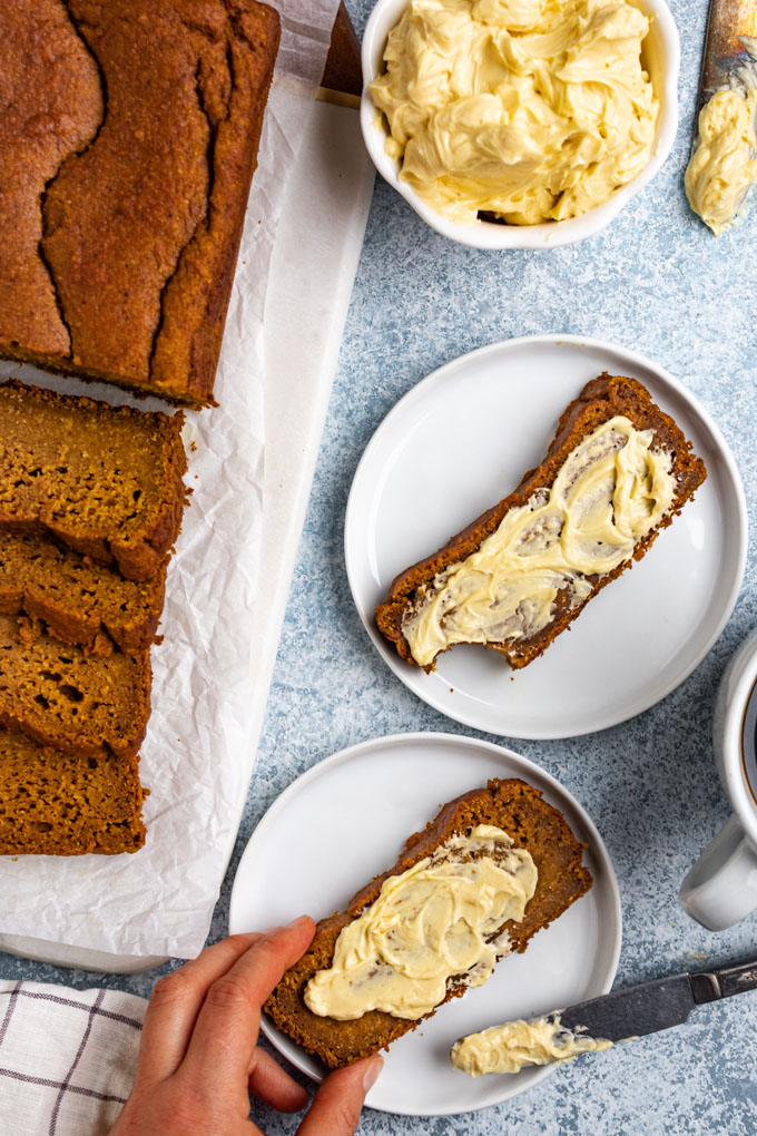 Healthy pumpkin bread on a light blue background. The pumpkin bread is partially slices, and off to the side. Two slices of pumpkin bread are centered., on white plates. They are spread with maple whipped butter, one of them is being held by a hand.