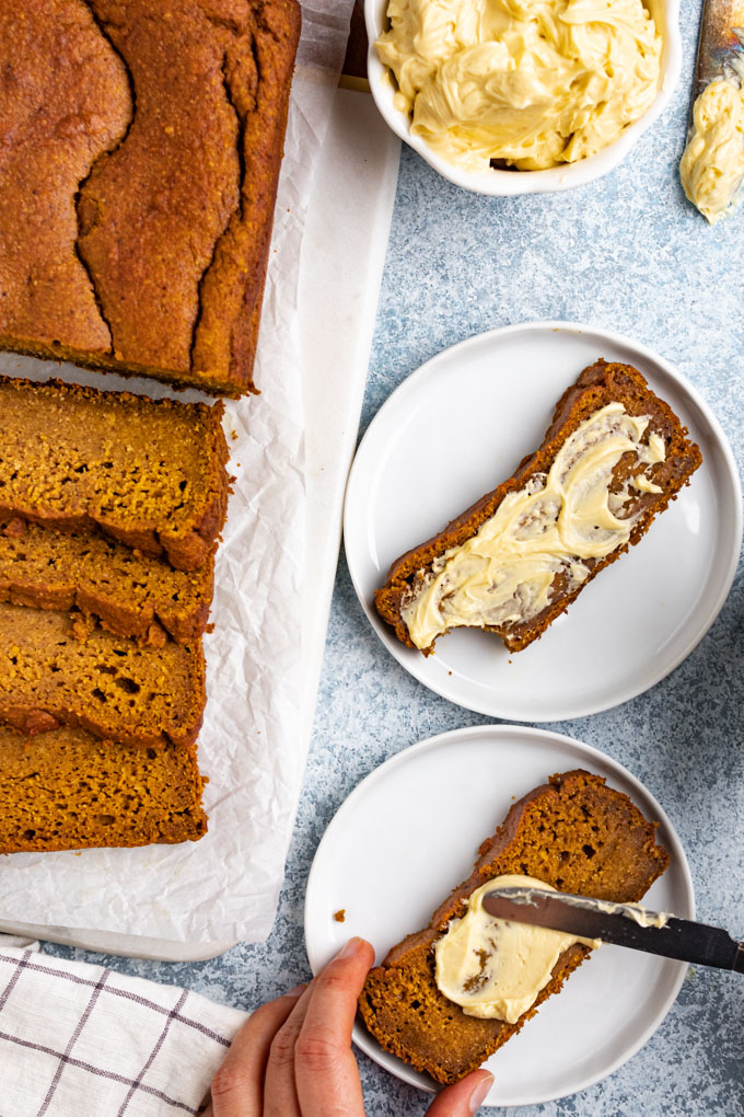Healthy pumpkin bread is sliced, on a white platter, off to the side. Two slices of pumpkin bread on white plates are in the center. One slice is being held by a hand and has knife spreading maple butter on it.