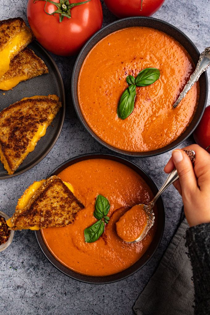 Overhead shot of roasted tomato and red pepper soup on a dark gray background with dark gray bowls.. A hand is spooning some of the soup up. There are basil garnishes on the soup. A half of a grilled cheeses sandwich is resting in the bowl of tomato soup.