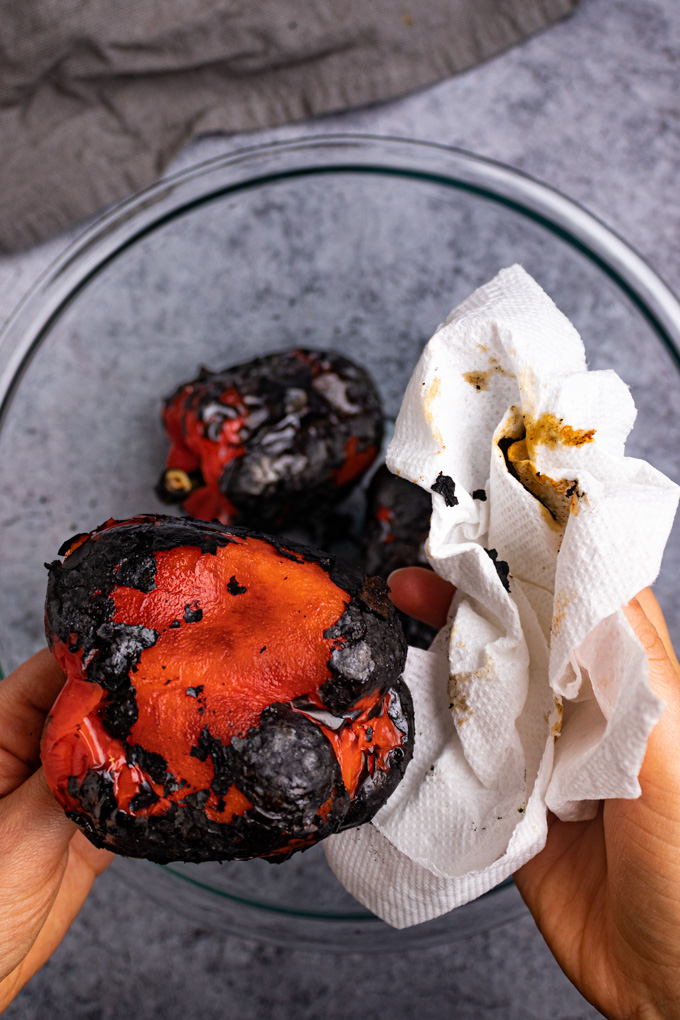 A roasted red pepper is being held by two hands, one hand is using a paper towel to peel away the charred skin.
