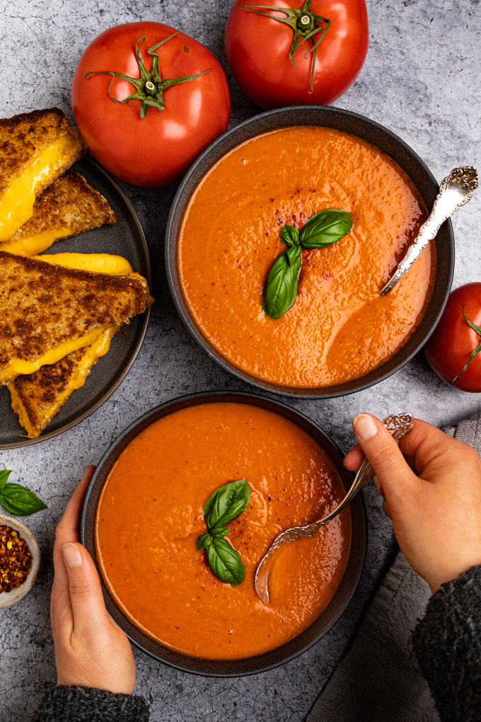 Overhead shot of roasted tomato and red pepper soup. Two dark gray bowls are filled with soup, the soup is garnished with basil leaves. A hand is holding a spoon in the soup, and another hand is holding the other side of the soup. A plate of grilled cheese sandwiches are in the background , a long with some tomatoes.