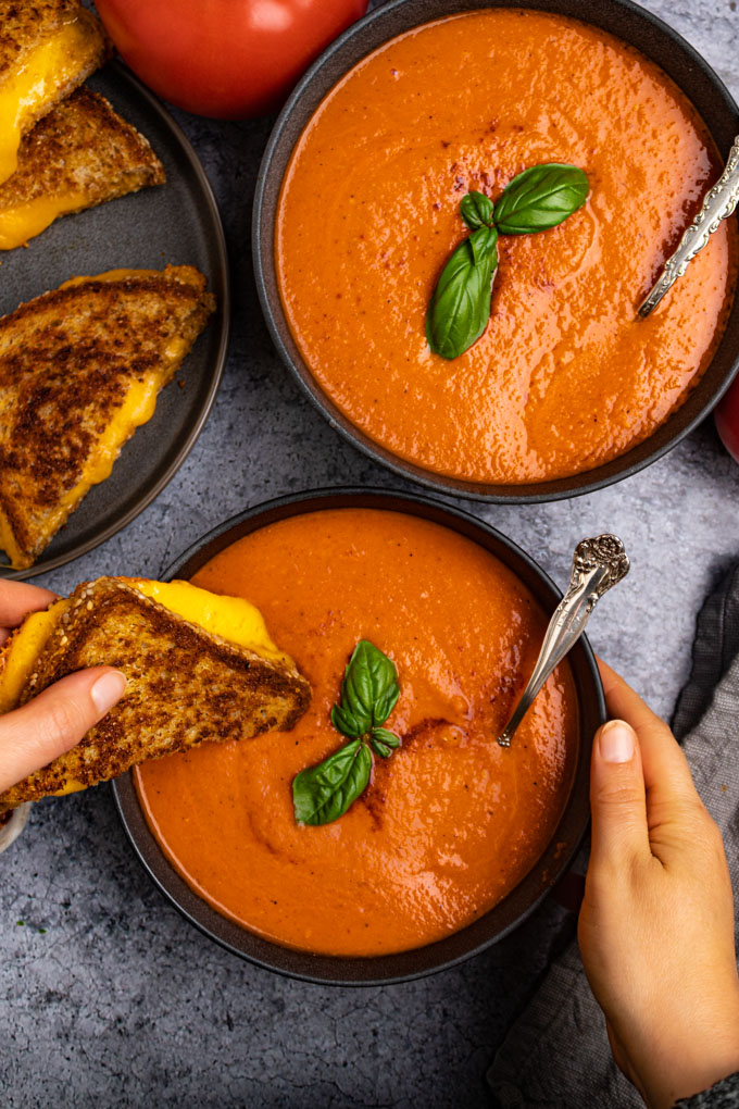 Overhead shot of roasted tomato and red pepper soup with a hand holding one side of the bowl, and the other hand is dipping a sliced of grilled cheese into the soup.