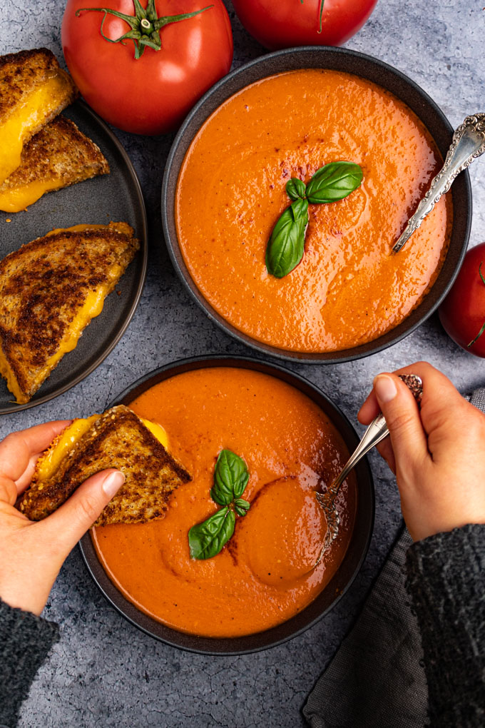 Overhead shot of roasted tomato and red pepper soup. One hand is spooning some of the soup, while the other hand is holding a slice of grilled cheese in the soup for dipping. A plate of grilled cheese sandwiches are in the background, along with some tomatoes.