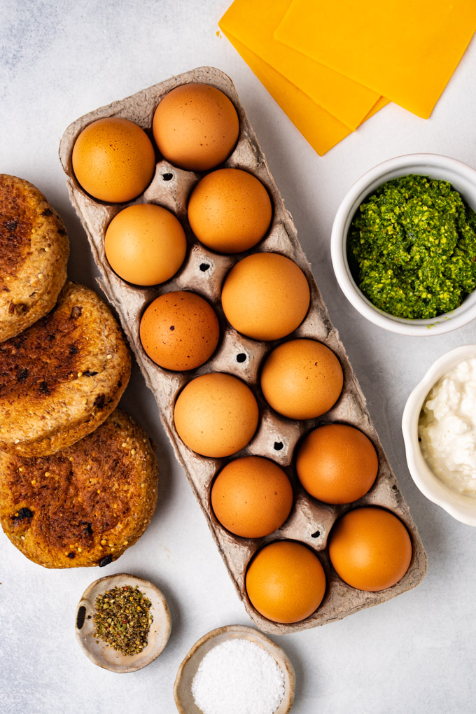 A carton of eggs, English muffins, cheese slices, salt, pepper, pesto, and cottage cheese.