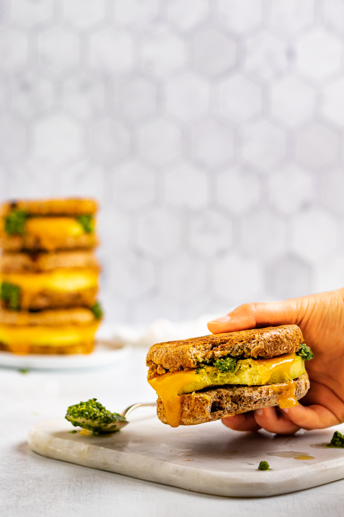 A close up shot of make ahead breakfast sandwiches with a hand holding the sandwich. A bite is taken out of the sandwich. A spoonful of pesto is next to the sandwich. A stack of 3 sandwiches are in the background.