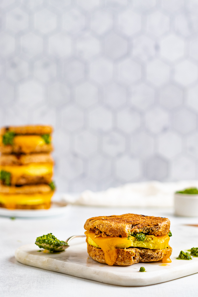 Straight on shot of a make ahead breakfast sandwich with pesto. A close up of the sandwich, with a bite taken out of it is sitting on a marble board. A stack of the sandwiches is in the background.