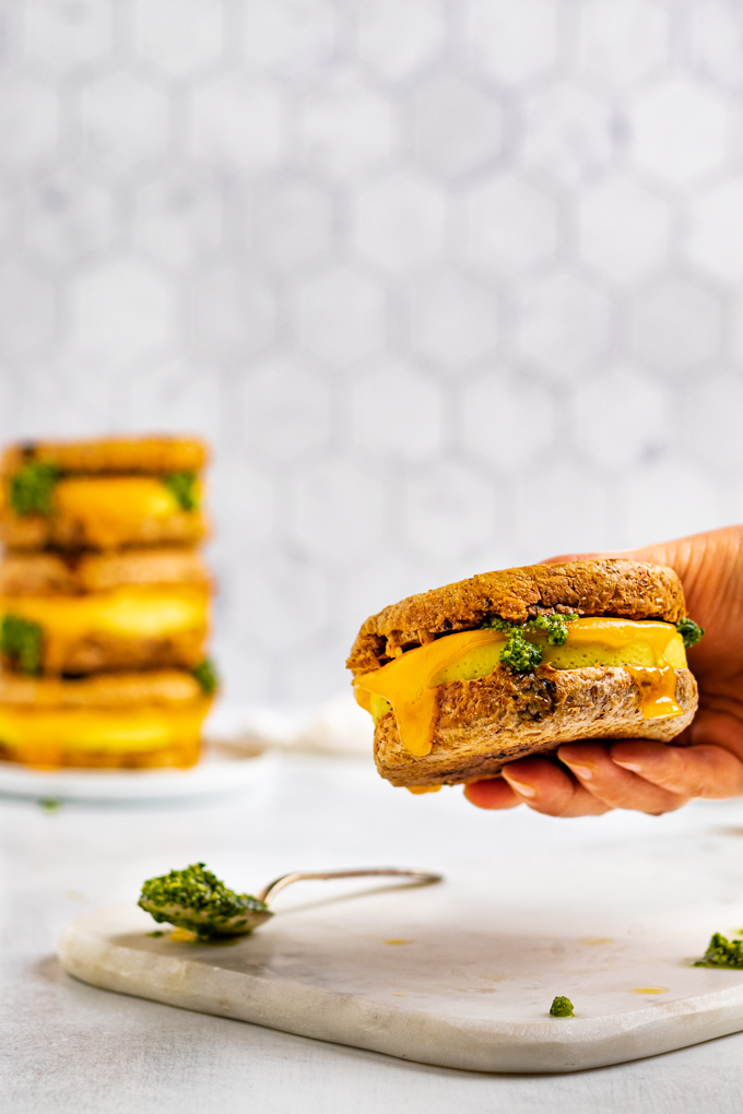 Closeup shot of make ahead breakfast sandwiches with pesto. A hand is holding the sandwich up in the air. A stack of 3 sandwiches is in the back ground. 