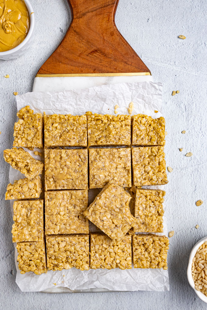 Healthy rice krispie treats are cut up into squares and lying on a marble and wood board with parchment paper underneath. There is a bowl of cashew butter of to the side, along with a bowl of rice cereal.
