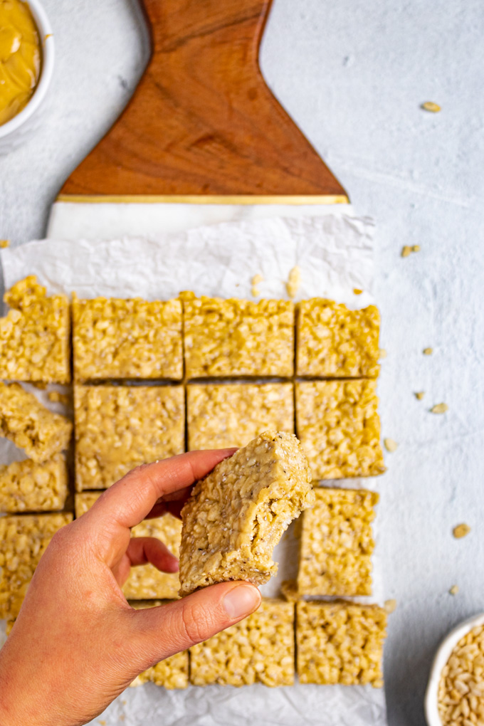 Close up shot of healthy rice krispie treats. The treats are cut into squares, and a hand is holding one of the bars, with a bite taken out of it. 