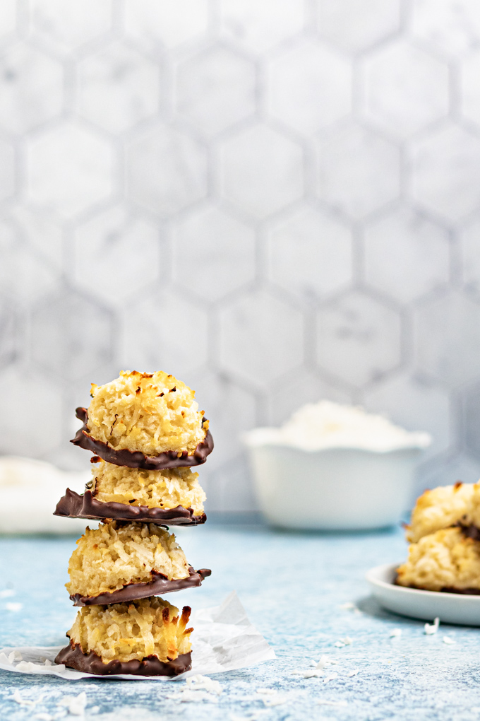A stack of 4 coconut macaroons on a light blue background. A plate of more macaroons is peaking in from the right.