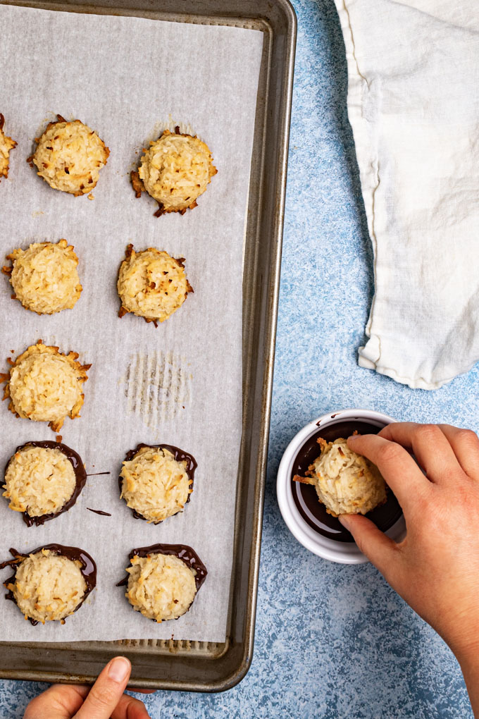 Baked coconut macaroons on a baking sheet. A hand is dipping one of the macaroons into dark chocolate.