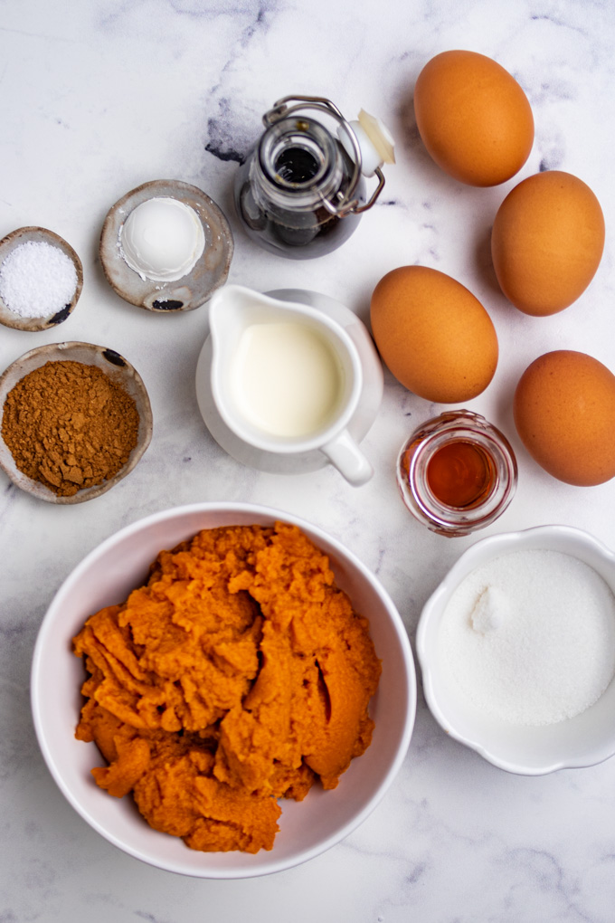 Ingredients are in bowls: pumpkin puree, granulated sugar, pumpkin pie spice, salt, heavy cream, maple syrup, and eggs