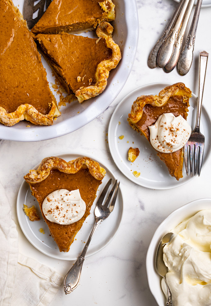 Overhead shot of easy pumpkin pie on a white marble background. Two slices of pumpkin pie are on small plates with dollops of whipped cream on top. A bowl of whipped cream is in the bottom right corner, off to the side. Extra plates, and forks are in the upper right corner, and the remaining pumpkin pie in a white pie plate are in the upper left corner.