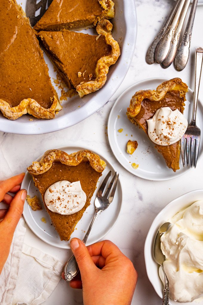 Overhead shot of easy pumpkin pie. 2 slices of the pie are on small plates, whipped cream is on top of each pie, and hands are holding the fork, and plate of one of the plates. There is a bowl of whipped cream in the lower corner, along with a pie plate with the remaining pie in the upper left corner. Extra forks are in the upper right corner.