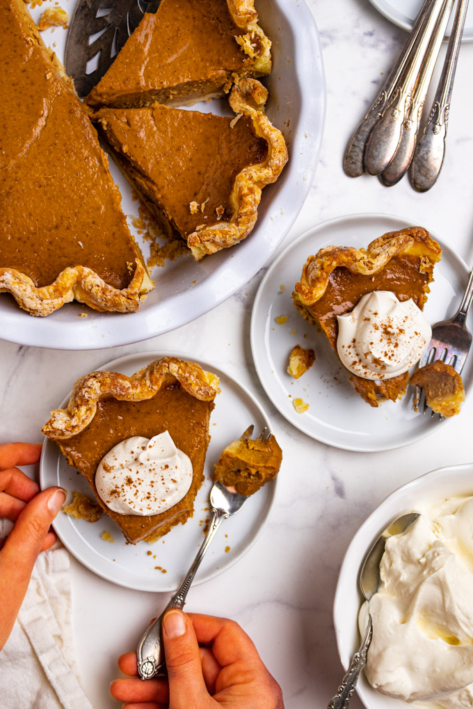 Overhead shot of easy pumpkin pie. Two slices of pie are on white plates with dollops of whipped cream on top. A bite has been taken out of each pie with a fork. Hands are holding the fork, and the plate. The remaining pie is in a white pie plate in the upper corner.