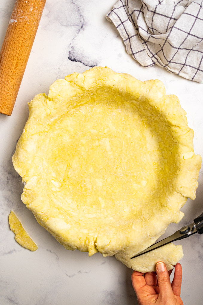 Gluten free pie crust is on a pie plate. A hand is using scissors to trim the excess dough.