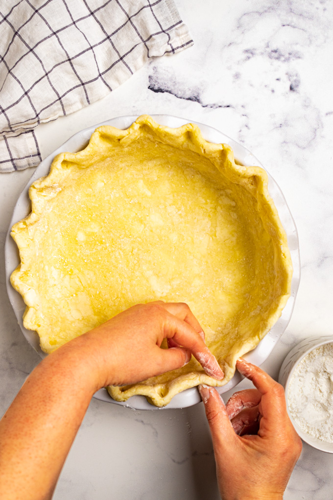 Hands are crimping gluten free pie crust in a white pie plate. There is flour in a small bowl to the side.