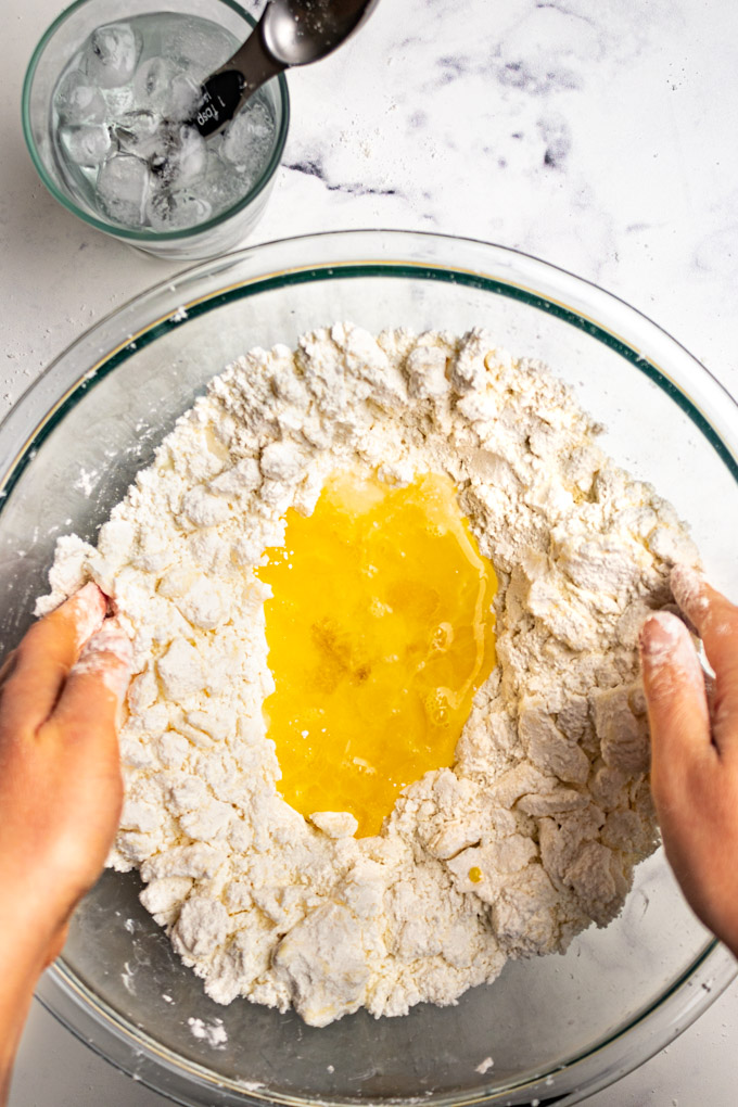 Egg, water, and vinegar being added to pie crust dry ingredients. Hands are reaching into the bowl about to toss things together.