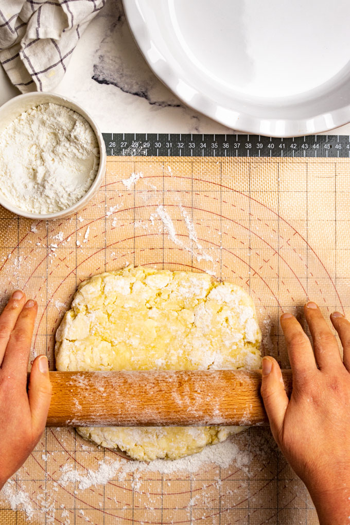Hands are using a wooden rolling pin to roll out gluten free pie crust on a pastry mat. A bowl of dusting flour is in the background.