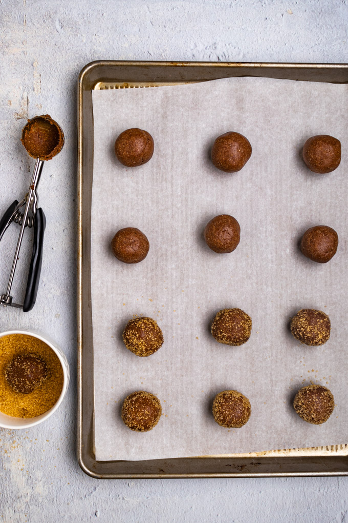 Balls of cookie dough on a parchment lined baking sheet. Some of the balls are rolled in demerara sugar. There is a cookie scoop off to the side, and a bowl of demerara sugar with a ball of dough in it.