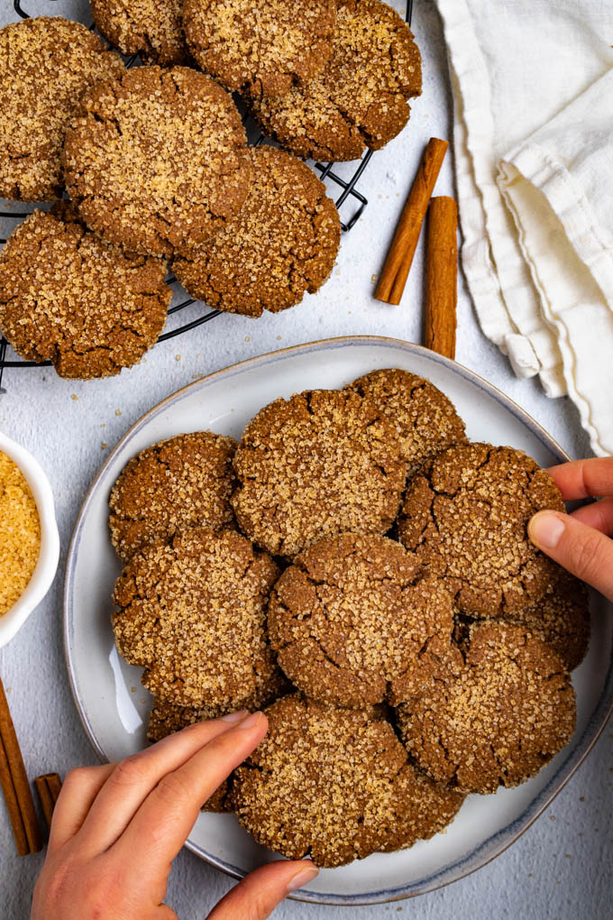 A plate of gluten free molasses cookies with two hands grabbing a cookie. More cookies are in the background on a round cooling rack.d