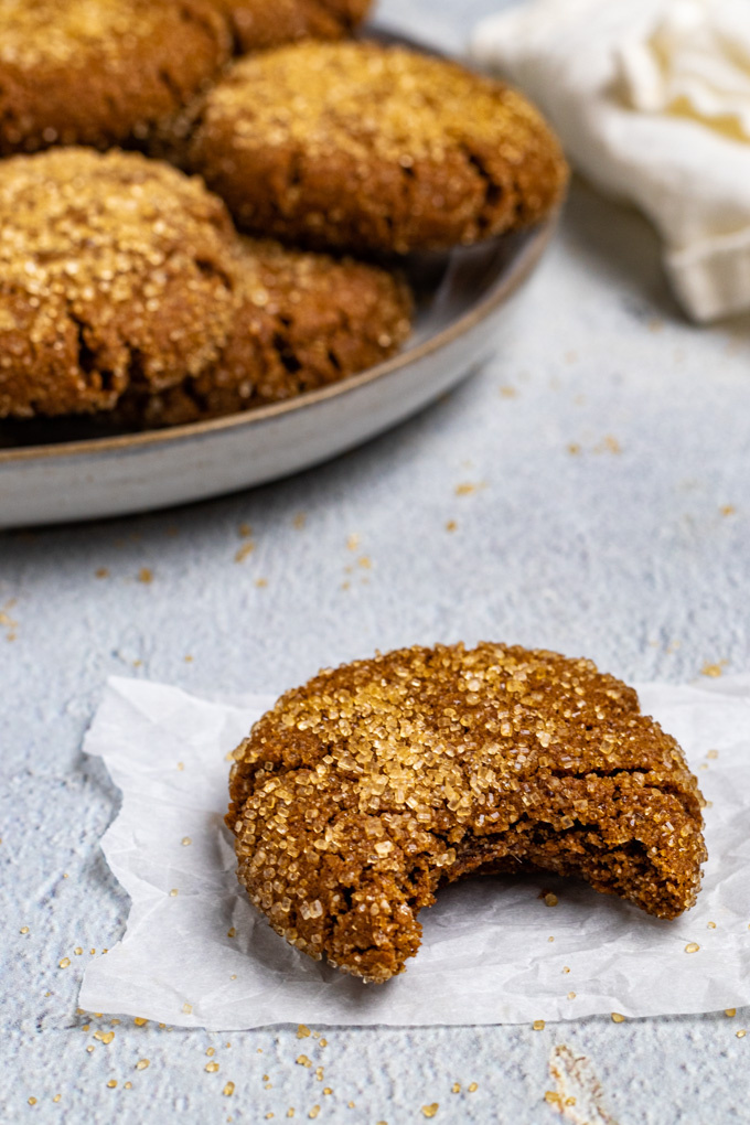 A close up of a gluten free molasses cookie with a bite taken out of it. The cookie is placed on a small piece of parchment paper. A plate of more cookies are in the background.