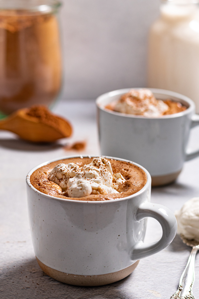 Angled, close up shot of a mug of homemade hot cocoa mix. The mug of hot cocoa is topped with whipped cream. Another mug is blurred in the background. There is a large jar of hot cocoa mix, and a jug of milk in the background..