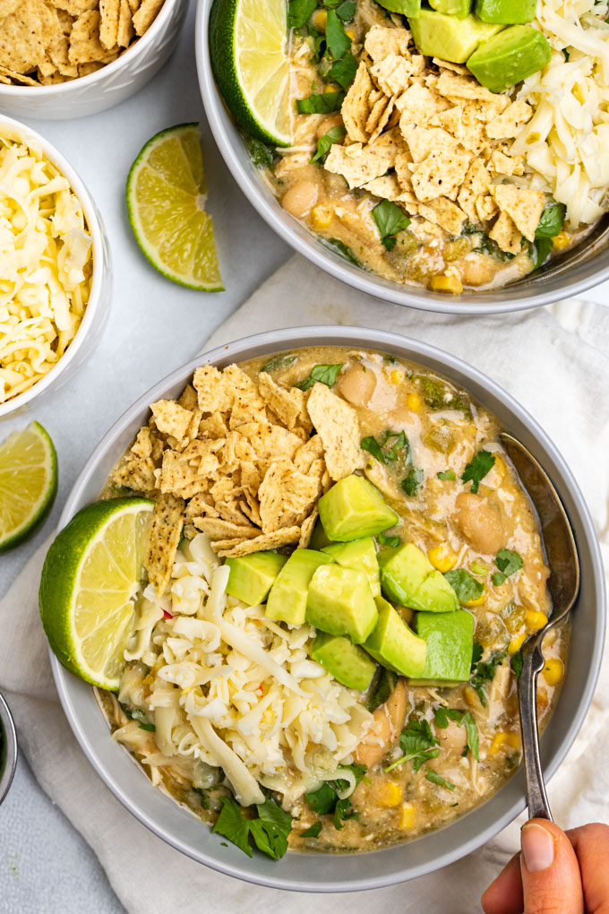 A bowl of white chicken chili with a spoon spooning some of the chili out. The chili is topped with avocado chunks, a lime wedge, shredded cheese, and crushed tortillas.