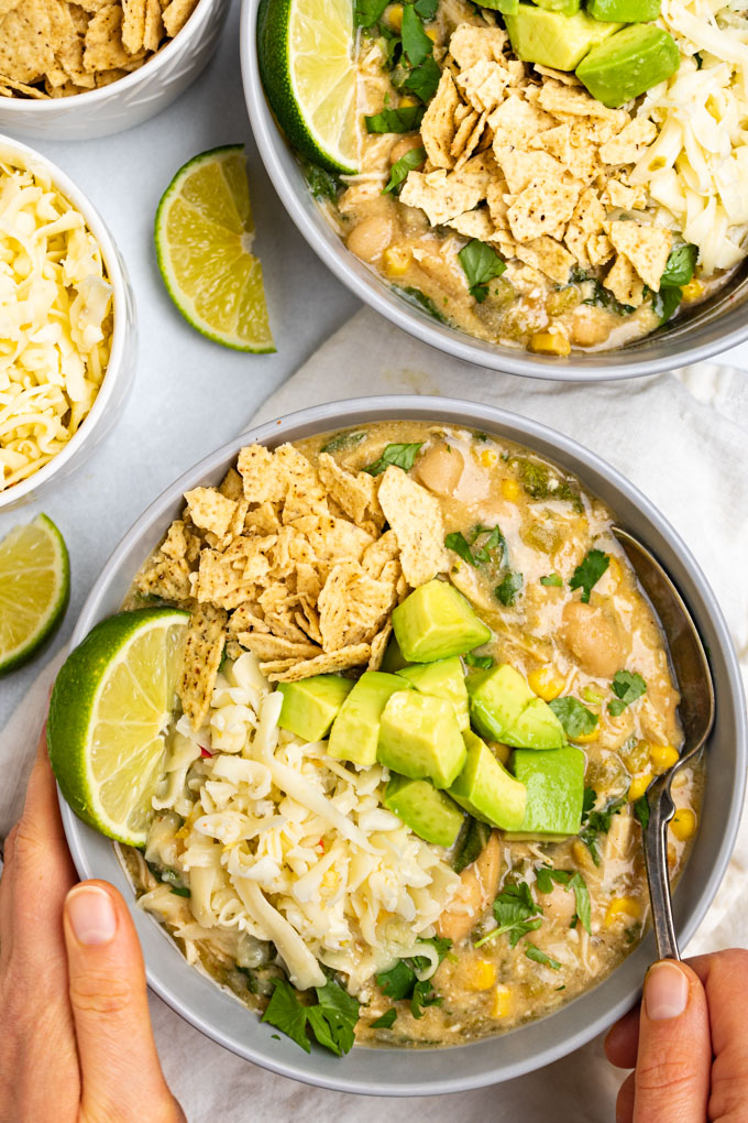 Overhead shot of instant pot white chicken chili. The bowl of chili is topped with avocado chunks, crushed tortilla chips, cheese, and a lime wedge. Hands are holding the bowl with a spoon in the chili.