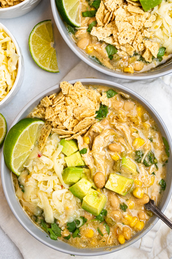 A bowl of white chicken chili with avocado chunks, shredded cheese, and crushed tortillas. The chili has been mixed up a bit with a spoon.