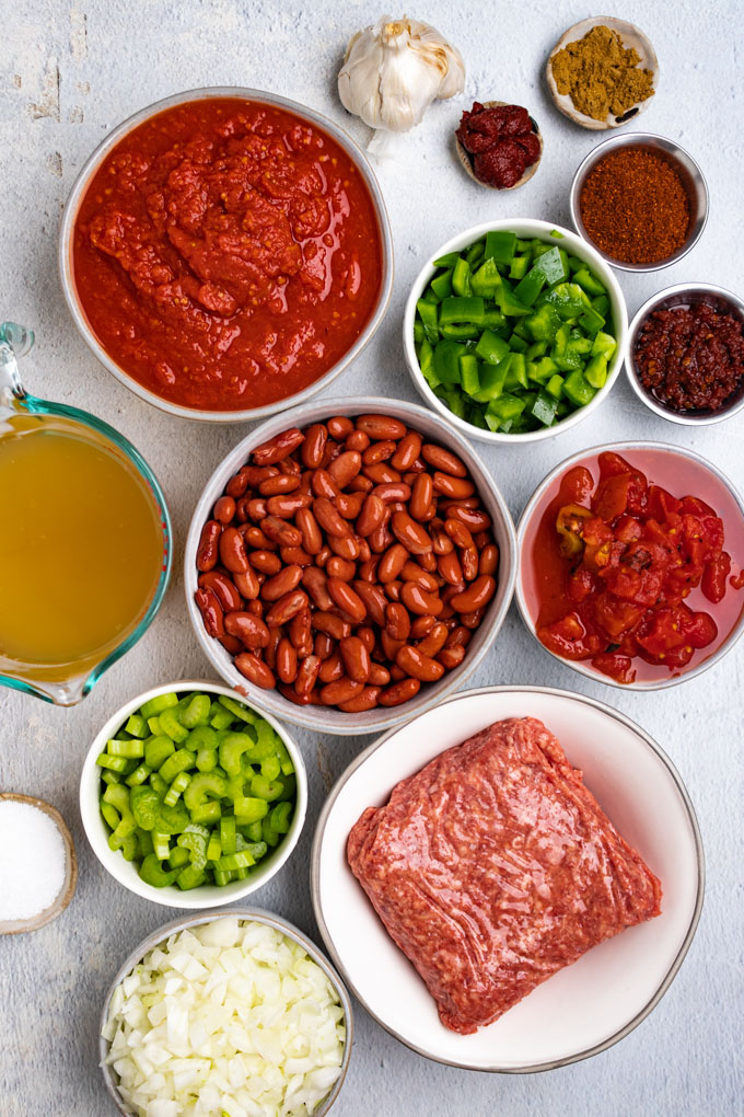 Ingredients for chili in bowls: red kidney beans, crushed tomatoes, green bell peppers, ground beef, roasted tomatoes, celery, onion, and broth.