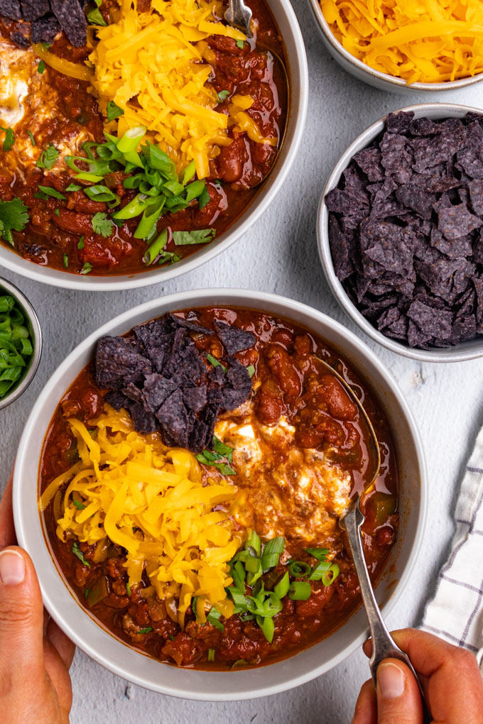Overhead shot of the best beef chili. The chili is topped with shredded cheddar cheese, sour cream, crushed tortilla chips, and green onion. A hand is holding the side of the bowl, while the other hand is holding a spoon.