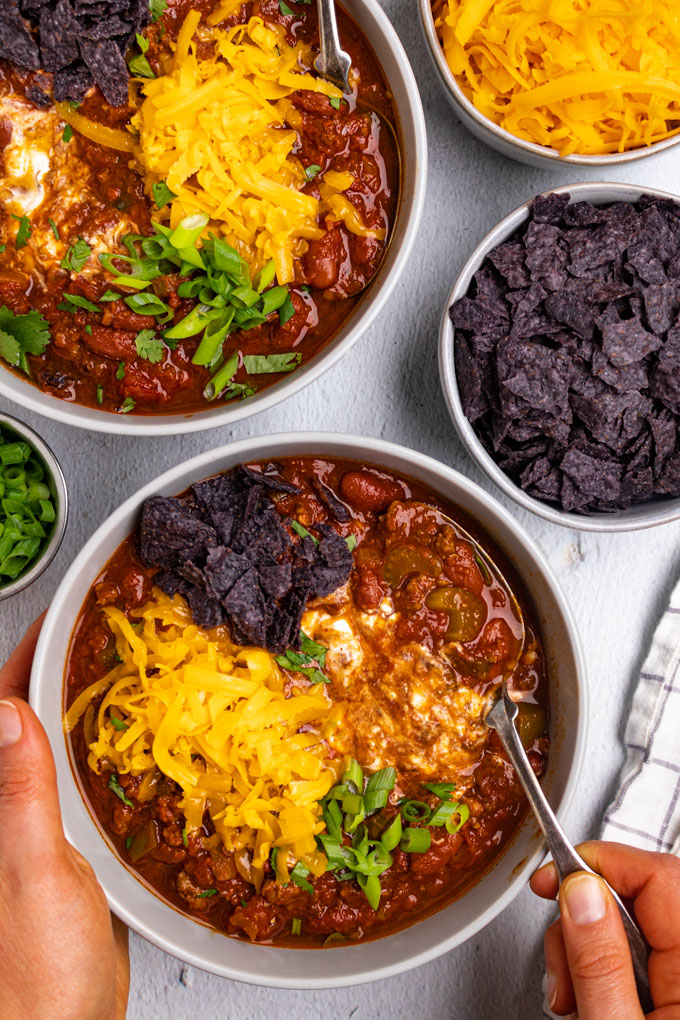 Overhead shot of the best beef chili. Hands are holding the bowl, and a spoon is spooning up the chili. Cheddar cheese toppings, sour cream, and crushed tortilla chips.