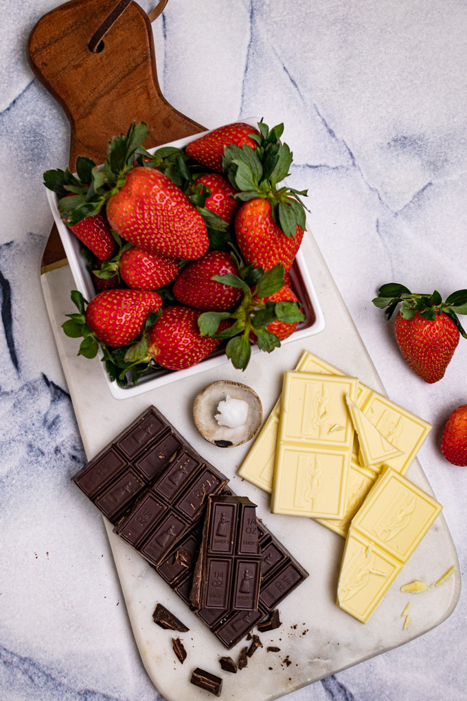 Ingredients are placed on a marble board - dark chocolate bar, white chocolate bar, coconut oil, and strawberries.