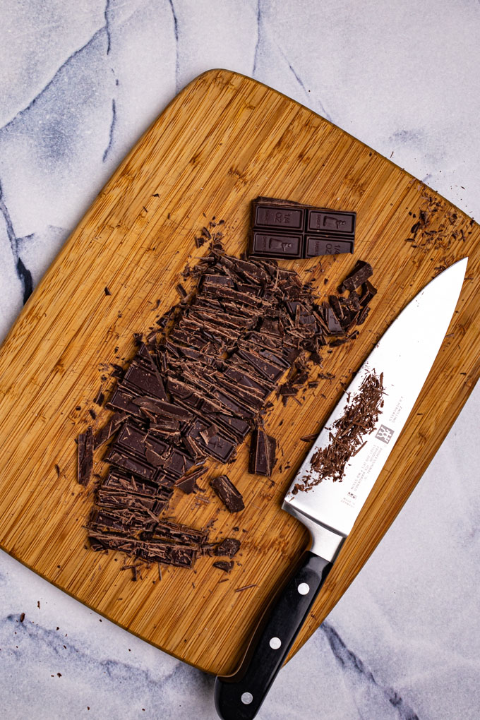 A dark chocolate bar has been chopped up on a cutting board. A chef's knife is laying beside the chocolate.