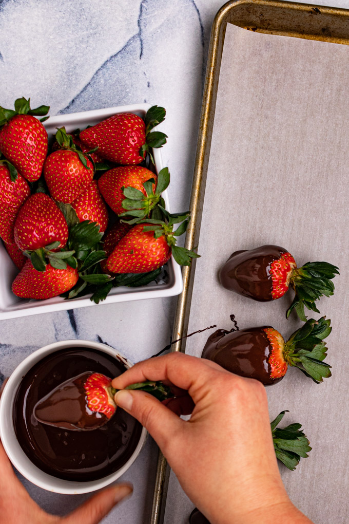 Strawberries are being dipped into a bowl of melted chocolate. Some are placed on a baking sheet lined with parchment paper.