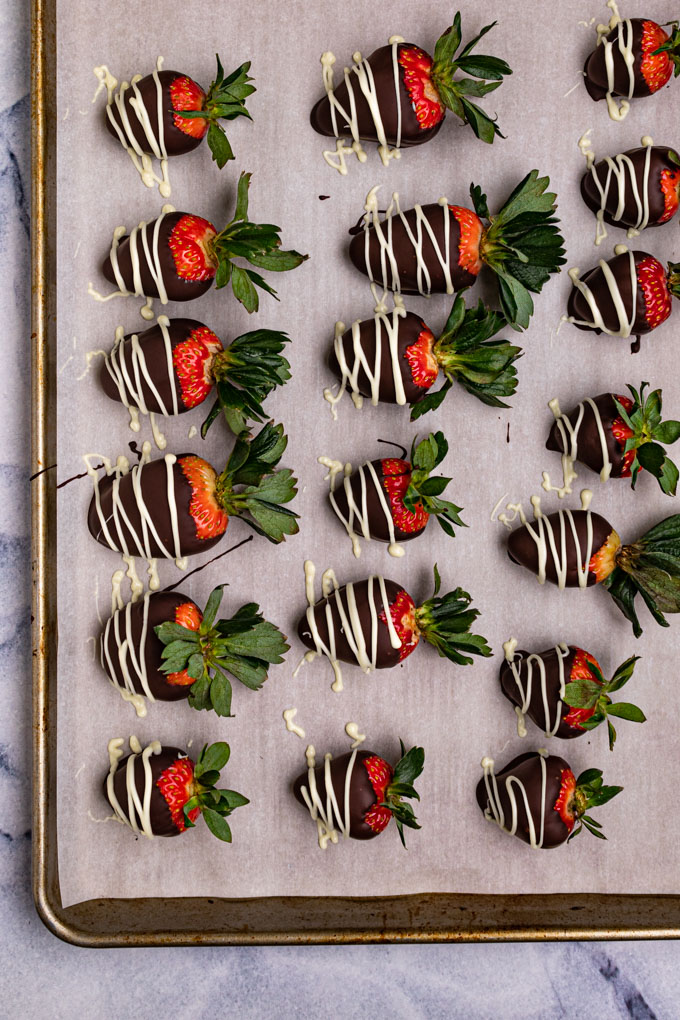 Chocolate covered strawberries are on a baking sheet lined with parchment paper. The chocolate is set up, and they have just been drizzled with white chocolate.