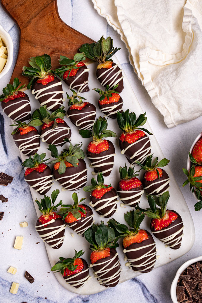 Chocolate covered strawberries are lined up on a marble board. Pieces are dark chocolate, and white chocolate are scattered around. There is a bowl of strawberries in the corner.
