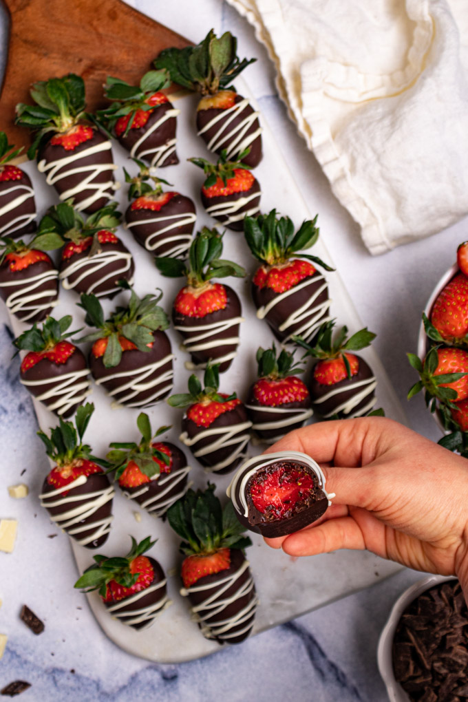 A hand is holding a chocolate covered strawberry that has a bite taken out of it. More strawberries are lined up in the background. 