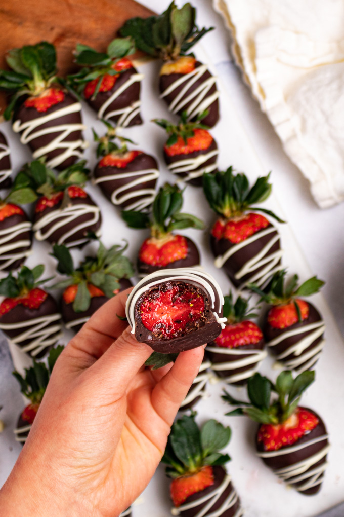 A close up shot of a hand holding a chocolate covered strawberry with a bite taken out of it. There are more strawberries underneath the strawberry with the bite taken out of it.