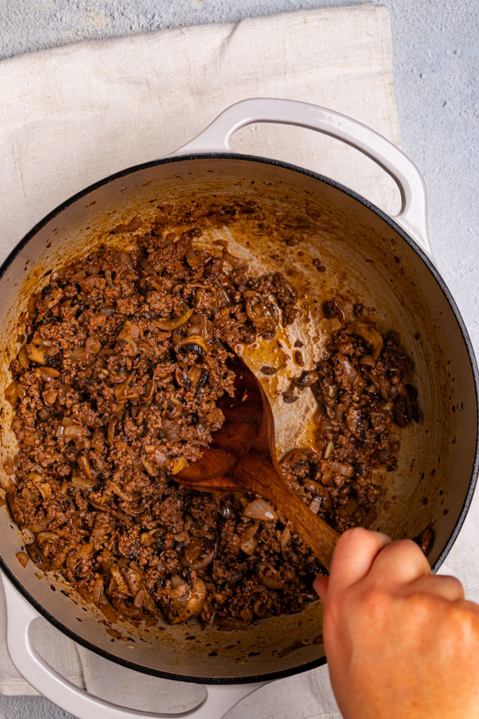 Overhead shot of ground beef in a dutch oven. A hand is holding a wooden spoon to stir the ground beef with.