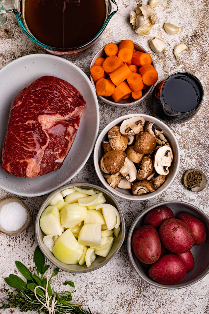 Ingredients for pot roast are in bowls - chuck roast, mushrooms, onions, carrots, red potatoes, herb bundle, garlic, beef broth, and red wine.